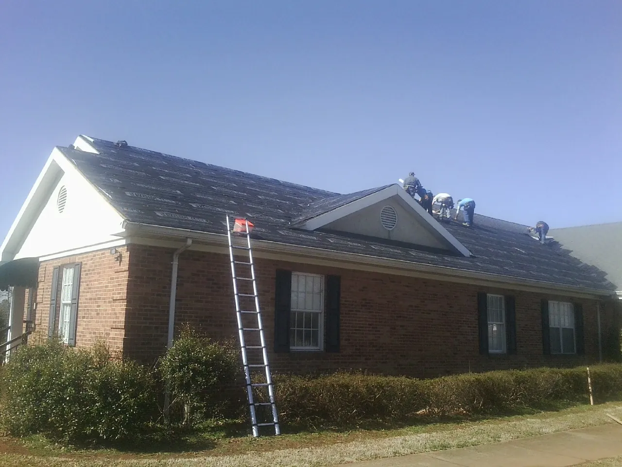 Crew tearing shingles off a brick ranch roof with a ladder set — Metal Roof Repair work by our crew in Davison